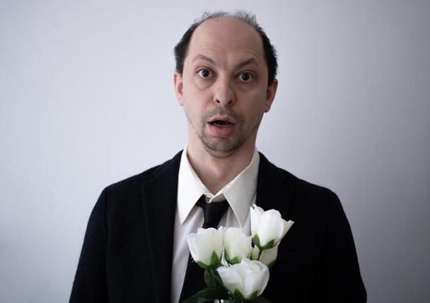 A balding man in a suit with a shocked expression holds white roses.