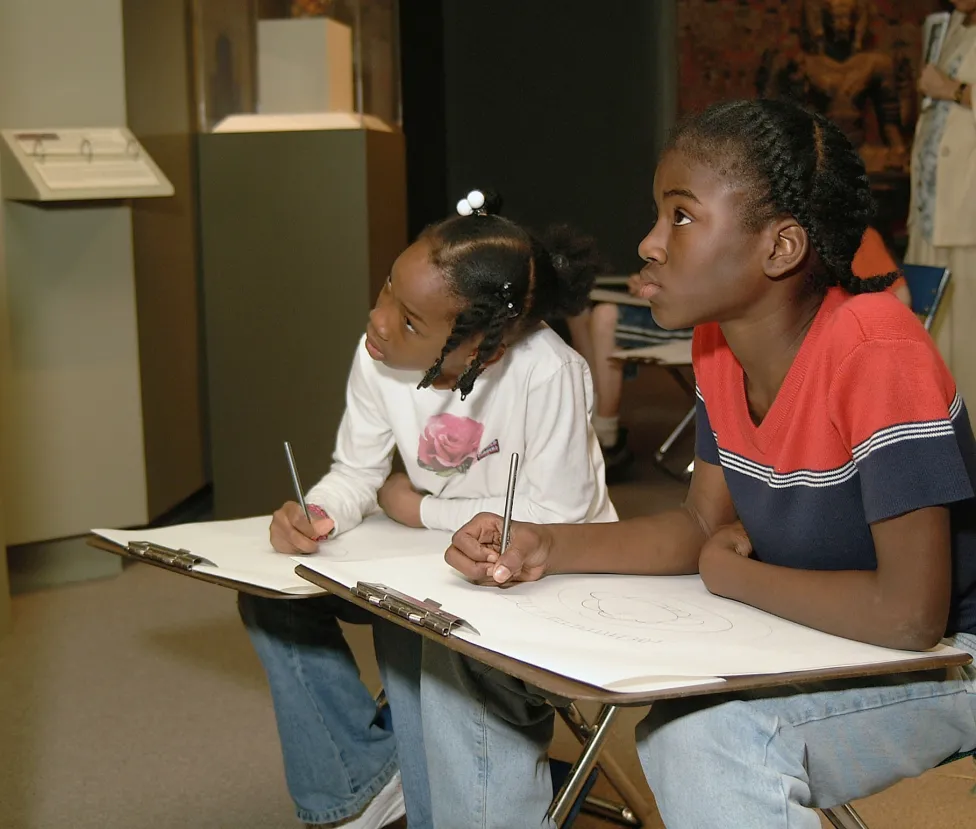Two children sitting in the DIA's Asian galleries and drawing on large clipboards.