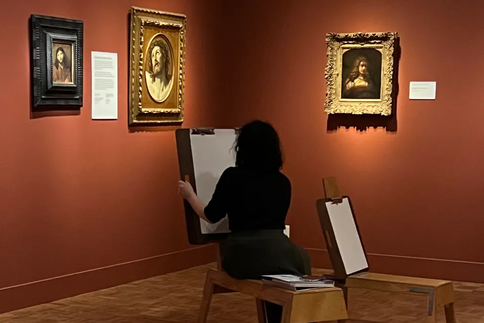 A visitor in all black sits on an easel bench drawing in the Dutch galleries