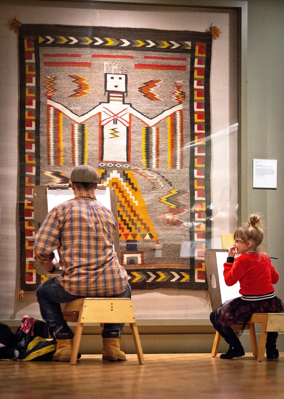 A man and a young girl sit on separate easel benches drawing in the Native American galleries