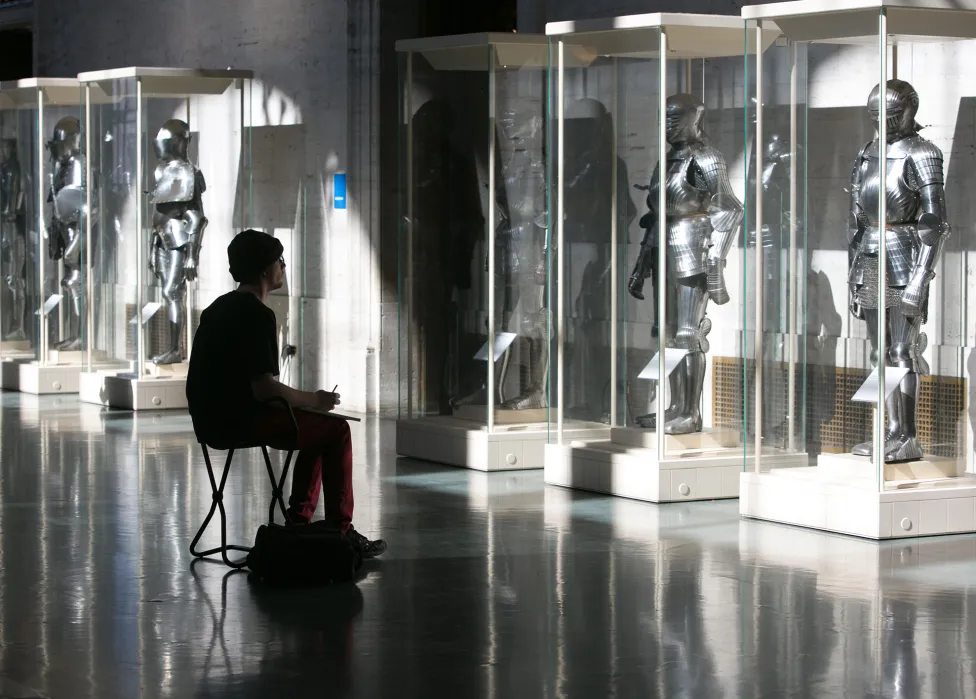 A patron sits drawing on a stool in front of suits of armor, shadowed from the light from the windows above the Detroit Institute of Art's Great Ha..