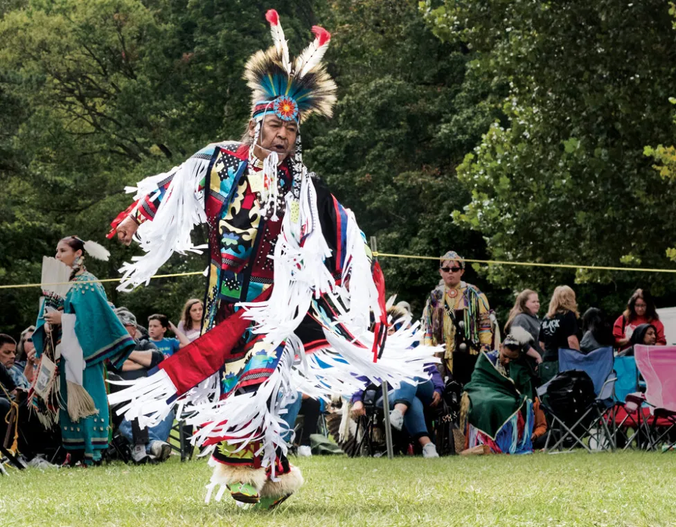 Reg Pettibone, dressed in Native American traditional clothing, dances on a green lawn with an audience behind him.