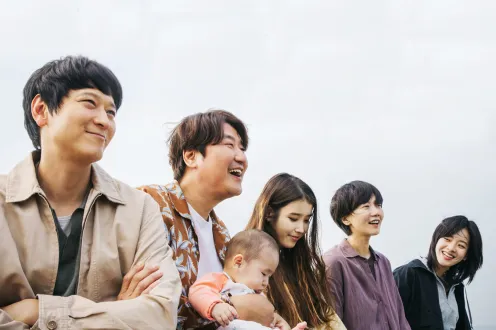 Two Korean men and three Korean women stand laughing in a row while one of the men holds a baby.