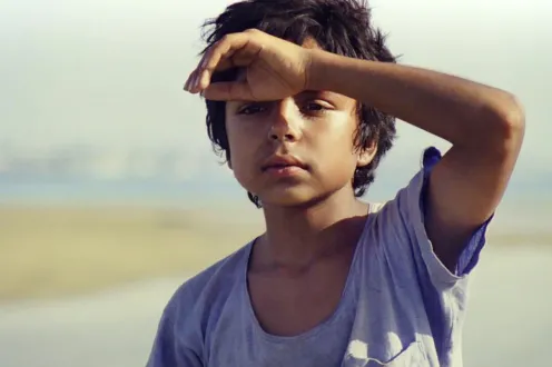 A young boy in a loose-neck t-shirt holds his hand up to his forehead as he looks out in the distance.