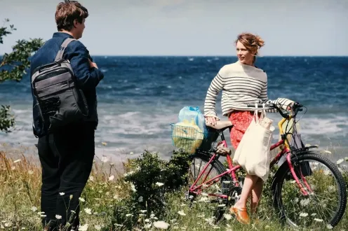 A woman sits on a bike in a flowery field in front of a beach, while speaking to a man with a backpack.
