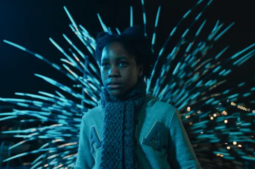 A Black girl wearing a sweatshirt and knit scarf stands alone with a smattering of lights behind her like fireworks.