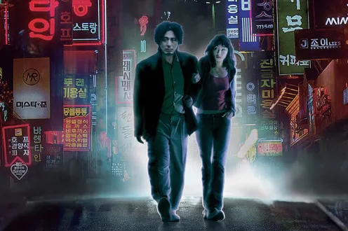A man and a woman with a ghostly air of light around them walk in front of a busy Korean street.