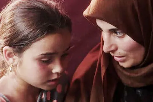 Two people keep their heads close together as they talk to one another. One is a younger girl and the other an older woman with a scarf around her head.