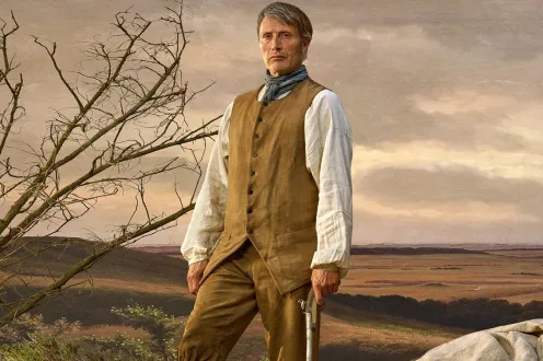 A man in 18th century clothing holds a gun while standing in an open plain.